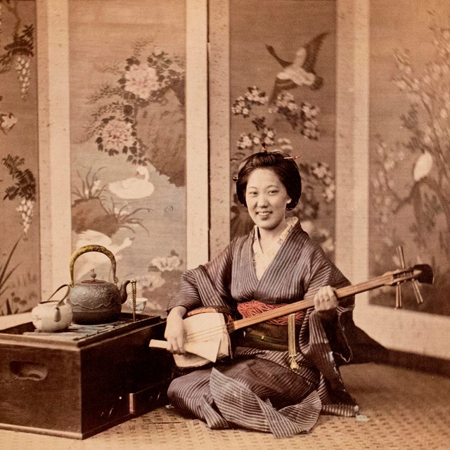 Hand-colored photograph of Japanese woman kneeling wearing blue kimono, holding instrument.