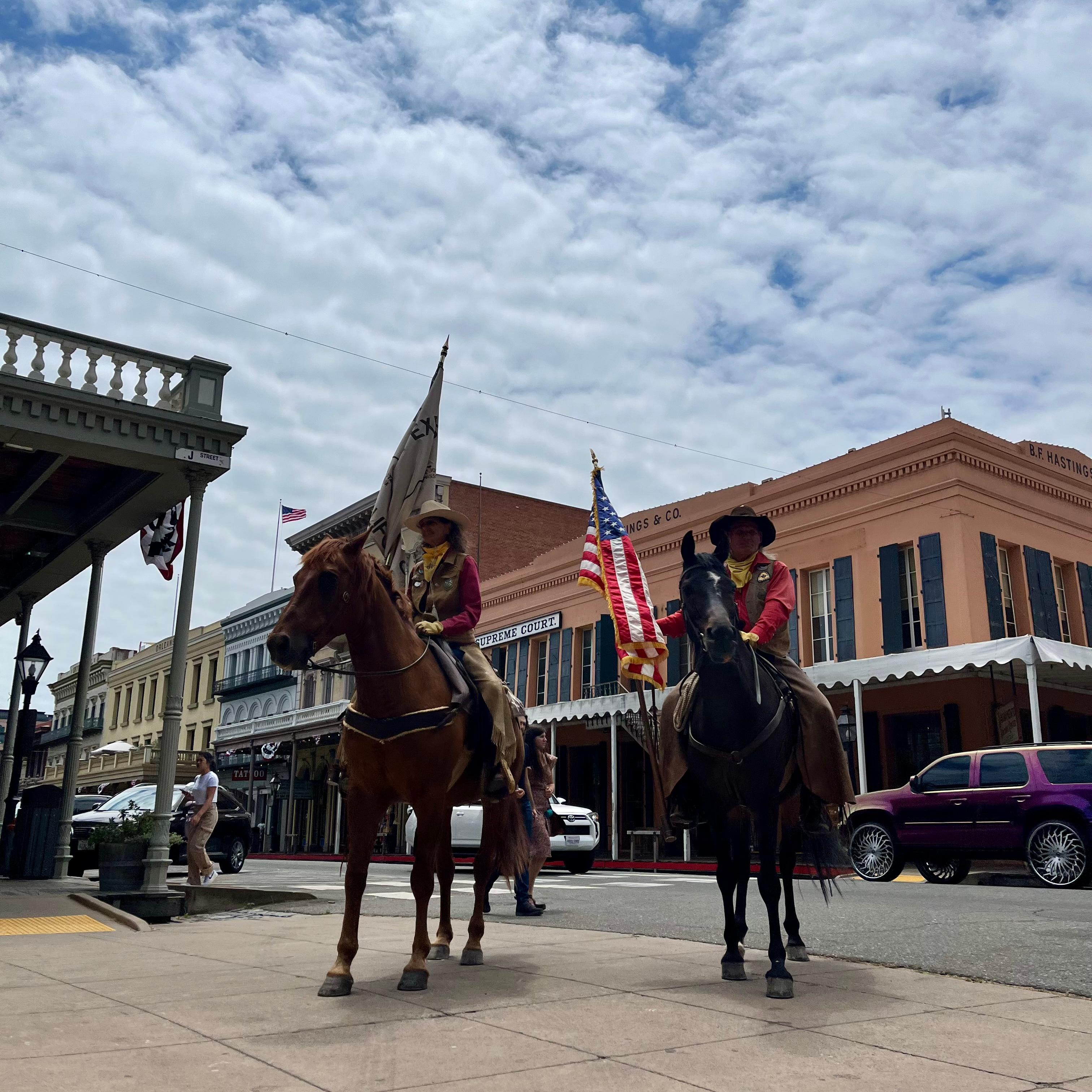 Two people in historic dress on horses, holding large flags.