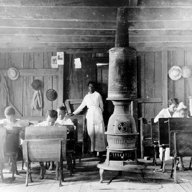 Several African American children in an old style school room with a wood stove in the middle.