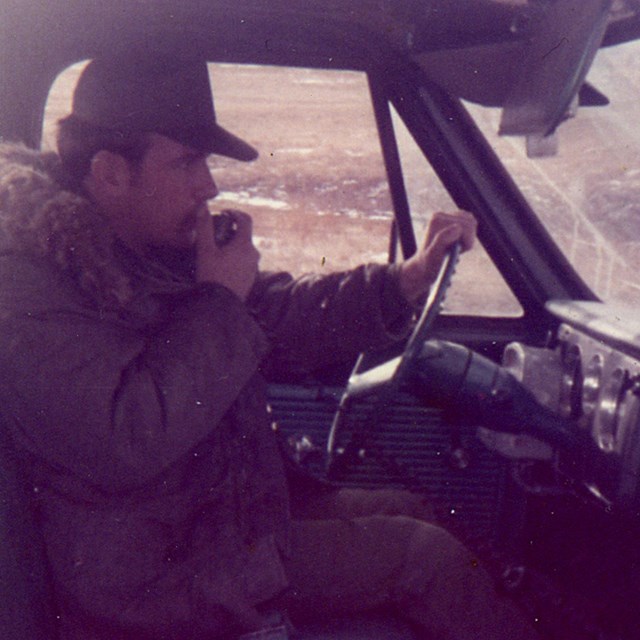 An airman, wearing a winter coat and cap, is seated in a car speaking into a vehicle radio