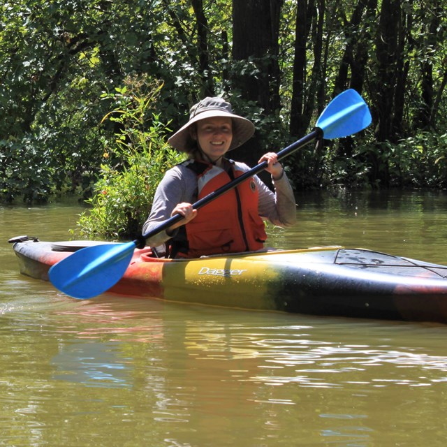 2 kayakers paddling on a sunny waterway through a wooded area