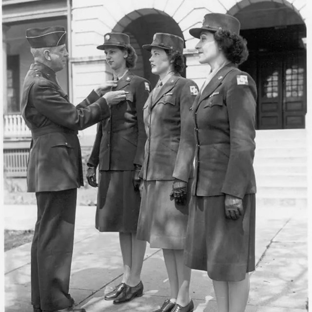 Three women in uniform face an older man in uniform who pins something onto one of their lapels