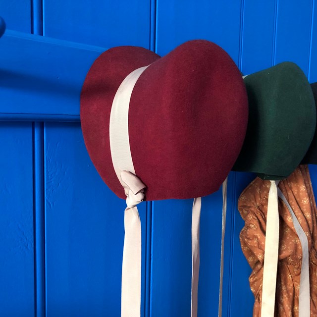 Red, green, and blue girls' 19th century-style bonnets hanging on pegs.