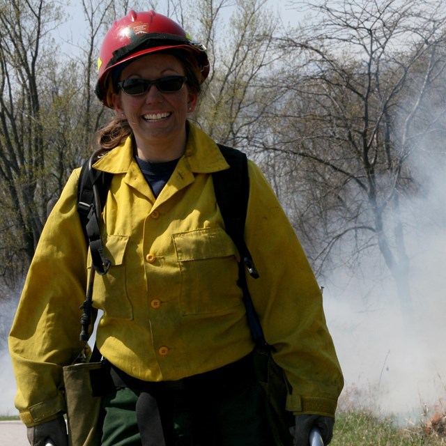 A woman in wildland fire protective gear with smoke in the background smiles at the camera.