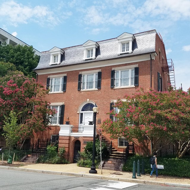 Photograph of brick three-story house. Blue, sunny sky in background.