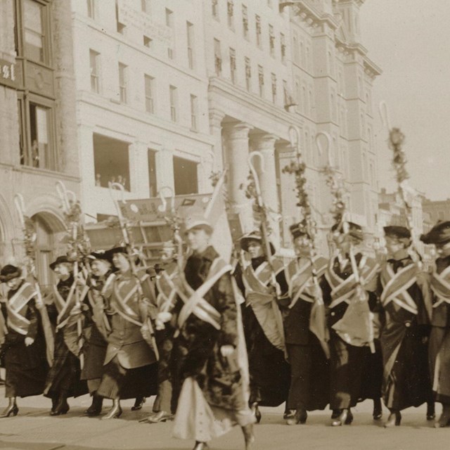Photograph of suffragists in sashes marching down the street past The Washington Post building, flan