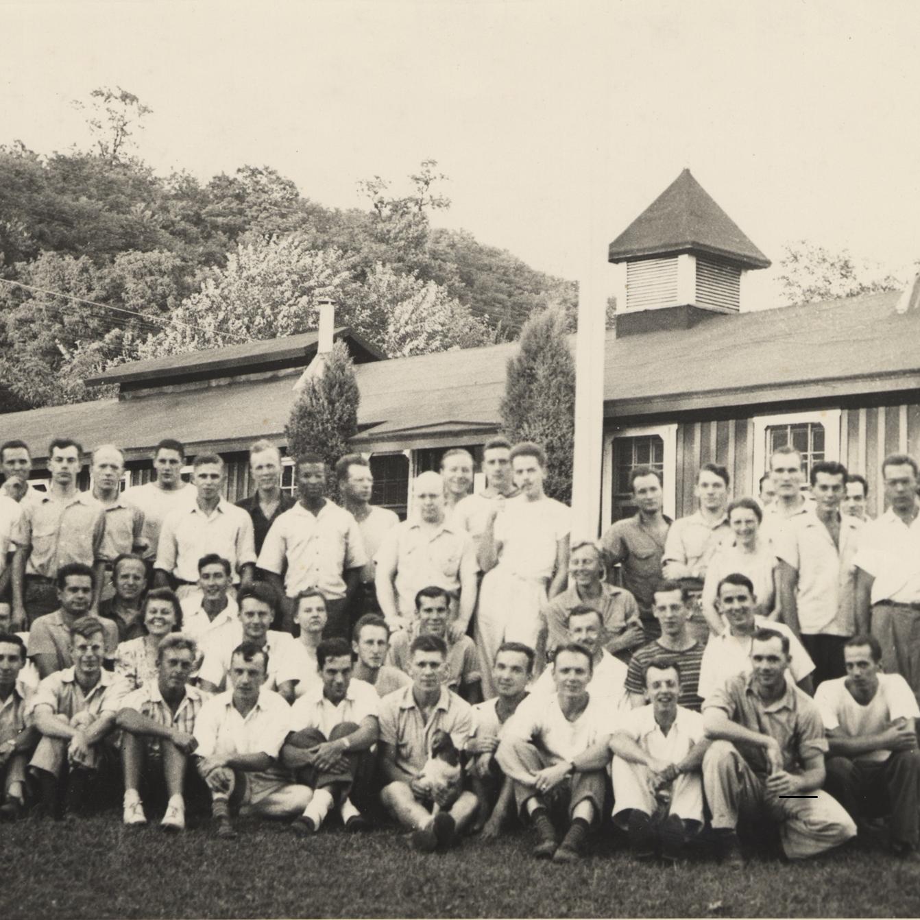 A group of young men smile in front of a low building. Most are white.