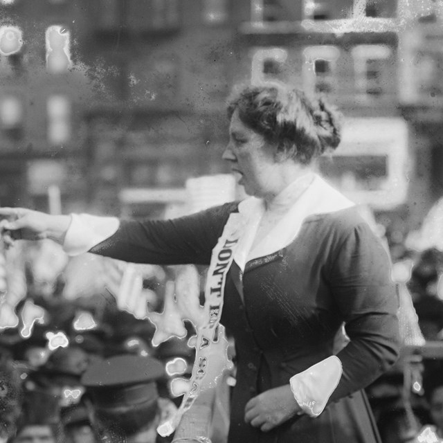 Margaret Hinchey wearing votes for women sash pointing at the crowd gathered around her