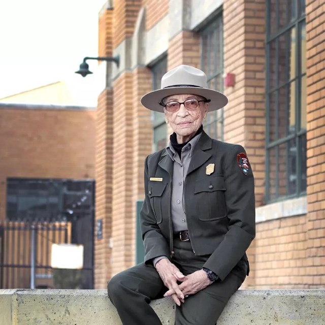 An older African American woman in park ranger uniform sits on low bench. 