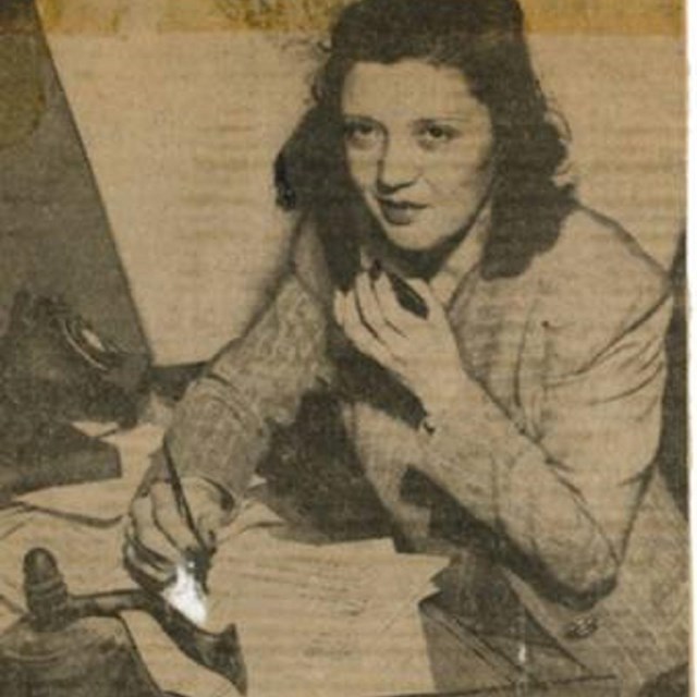 A young woman sits in front of a desk with papers while speaking into a phone receiver