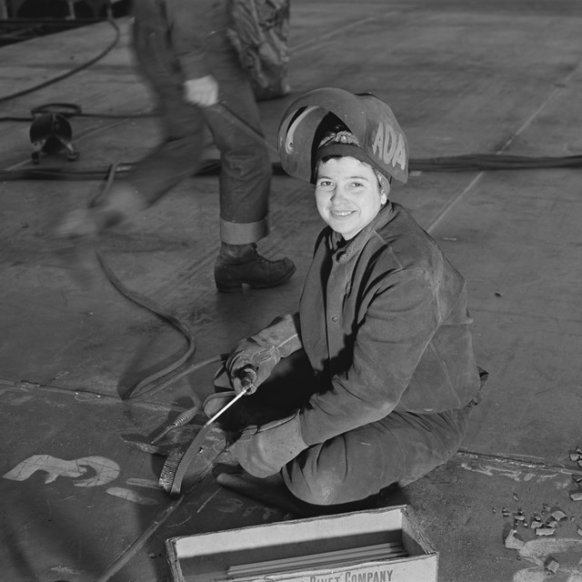 A woman in welding gear smiles at the camera while sitting on concrete floor