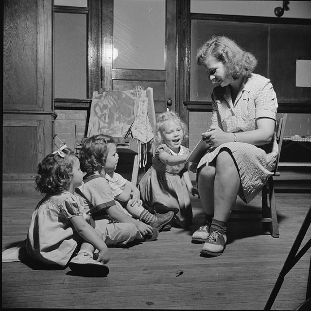 A young woman sits in a low chair and reads a book to three young girls