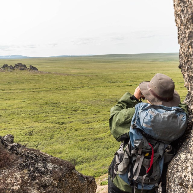 A backpacker looks at granite tors across the tundra through binoculars.