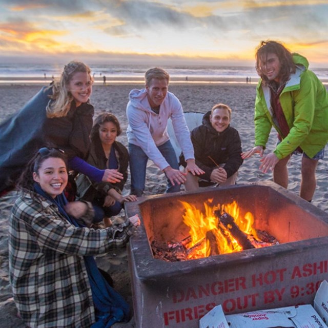 Group of men and women gathered around a fire pit on Ocean Beach at sunset