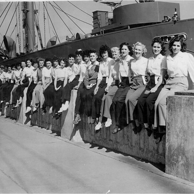 A row of women site on a wall in a shipyard, with a large ship in the background, black and white