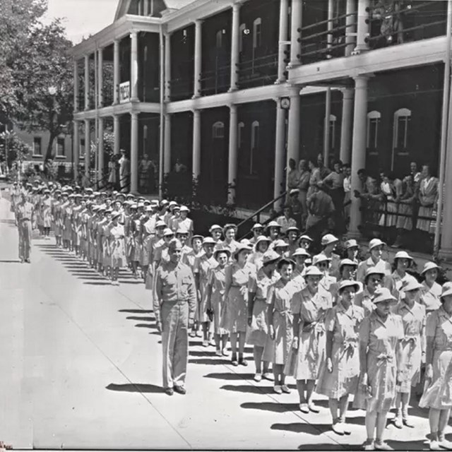 Women in uniforms walk down a street in straight lines while others look on 