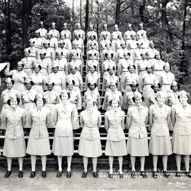Women in light colored uniforms smile at the camera standing in straight rows on bleachers