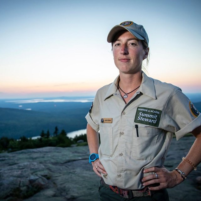 Alanna stands on the top of a mountain in Acadia, the sun setting behind her