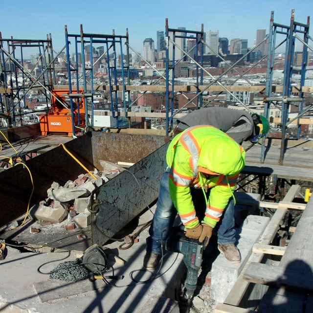 A worker in high visibility outerwear and blue jeans operates a small electric demolition hammer