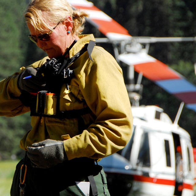 A women in protective gear and flight gloves speaks on a radio with a helicopter in the background.