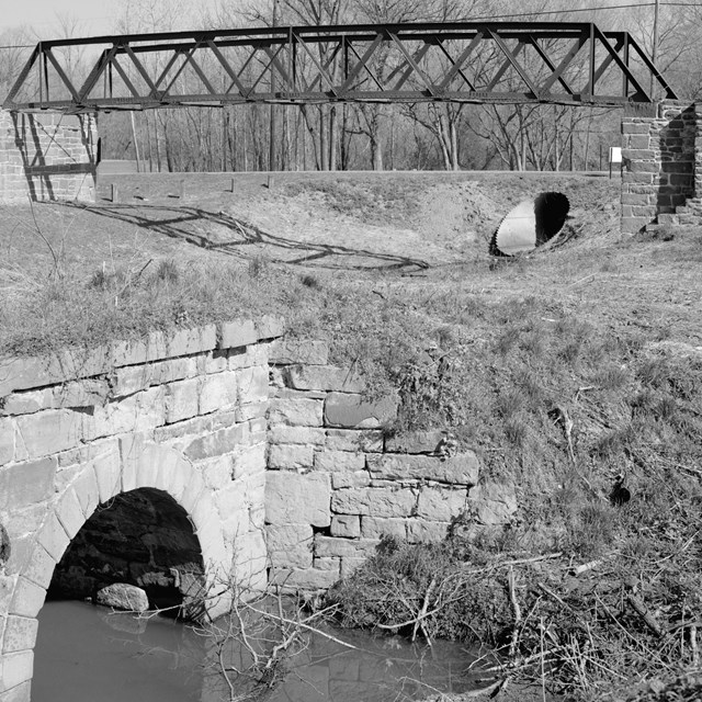 Greyscale photo of iron footbridge with stone culvert in foreground