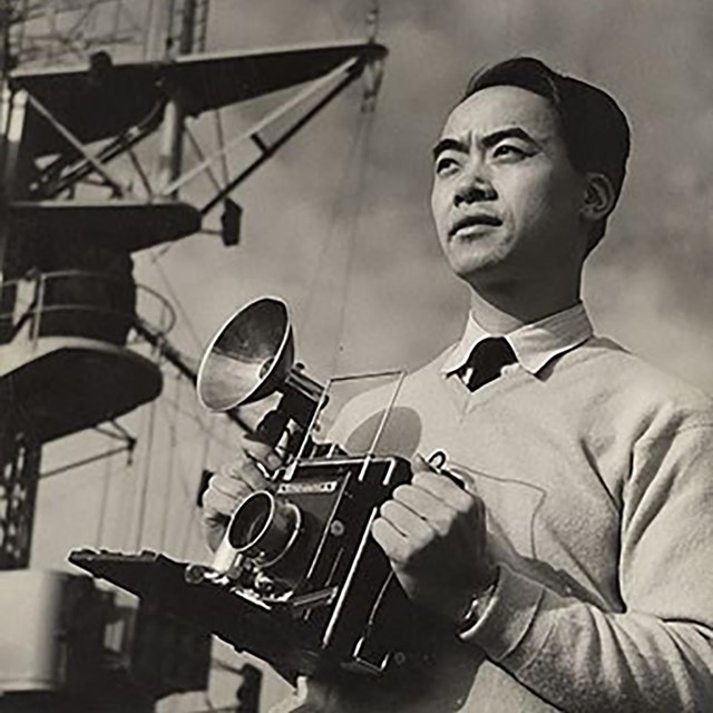 Black and white photo of an Asian American man holding a camera.