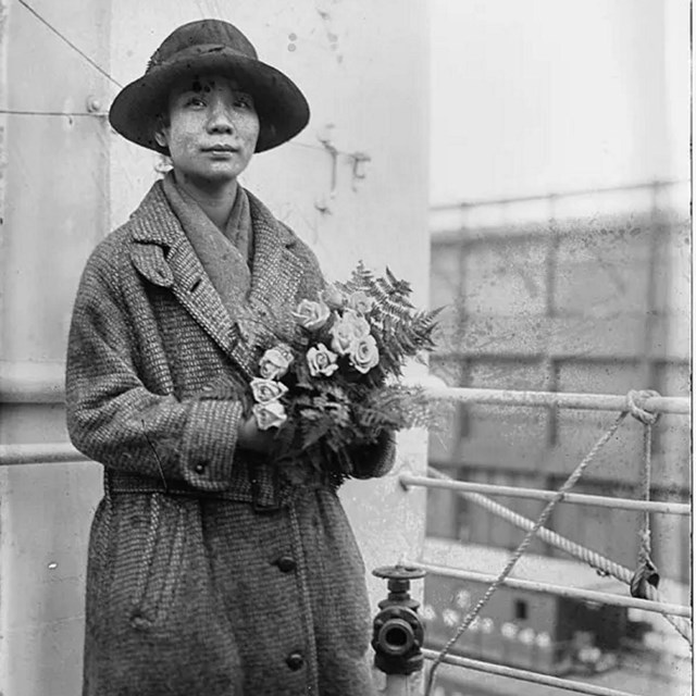 Woman with a broad brimmed hat and long coat stands alone looking at camera