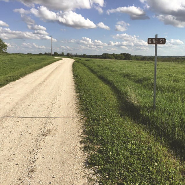 A gravel road though a prairie landscape goes by a sign labeled 
