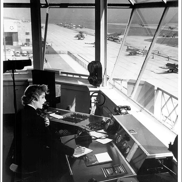 Woman speaking into microphone inside building overlooking runway and planes