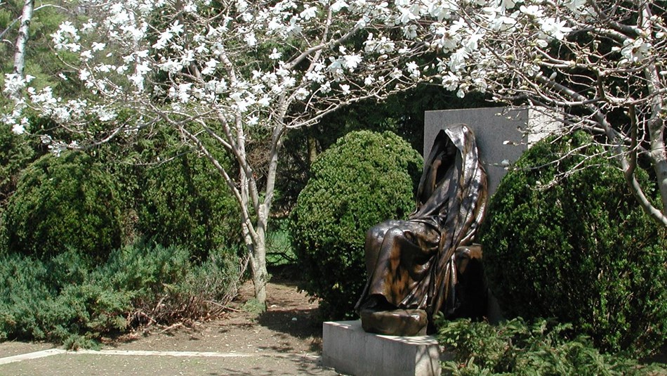 Bronze statue, in a garden framed by a flowering tree, of a cloaked woman