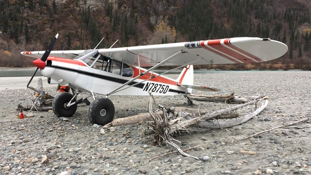 A fixed-wing aircraft with large wheels sits on a gravel beach near the water.