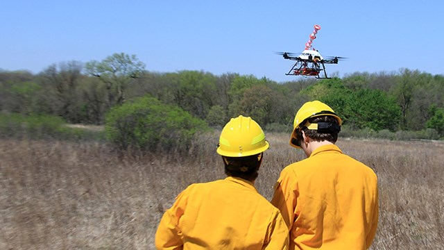 A man holds the controls for a stationary UAS while another man stand nearby.