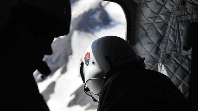 A crew member wearing a helmet peers out of a helicopter down on a mountain