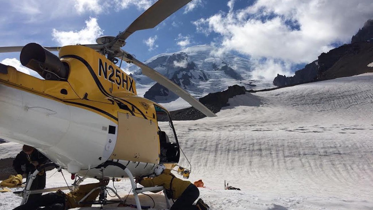 A man in a yellow shirt works under a helicopter parked on snow
