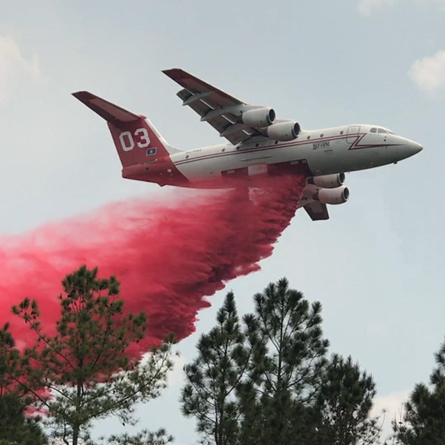 A large white airplane with red tail flies above the trees while dropping red fire retardant
