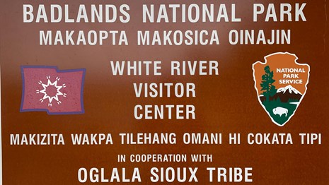 A sign with NPS logo and flag with tipis arranged circularly with greetings in English and Lakota.