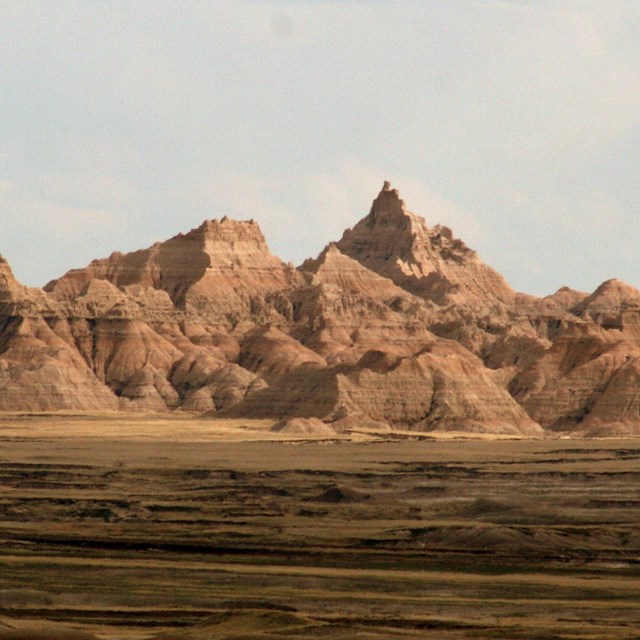layered badlands formation in distance under blue sky with green fields below.