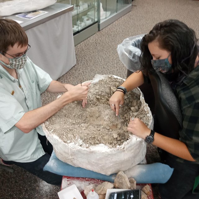 Two people in white lab coats crouch aside large round object.