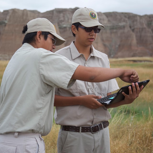 two volunteers in khaki uniforms use digital equipment in front of badlands buttes.