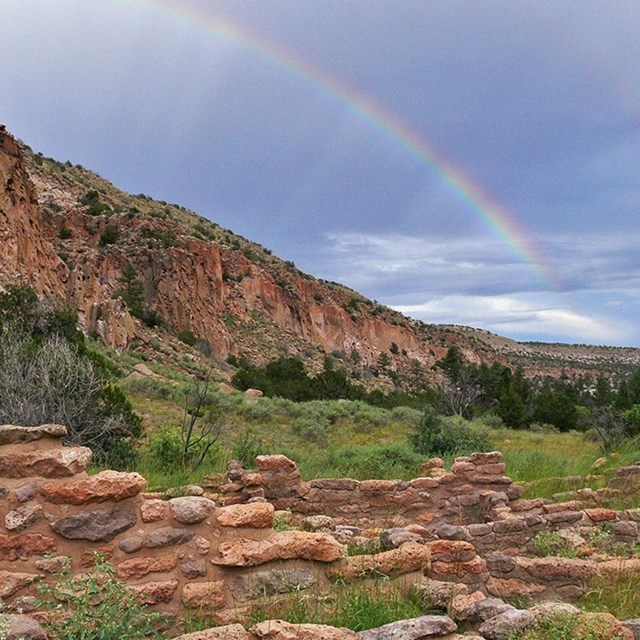 A rainbow in the sky above a canyon with the foundations of a building in the foreground.