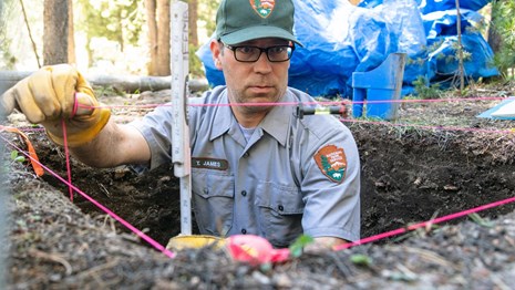 Man in NPS uniform stands in pit, measuring depth with string and ruler