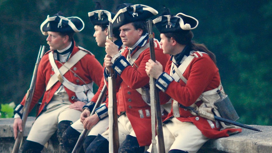 Redcoat soldiers sit on stone bridge holding muskets