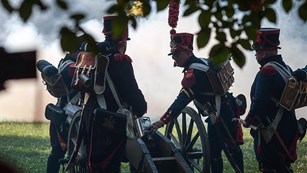 Soldiers in red and blue uniforms with boxy hats with large feathers confer around cannon in field