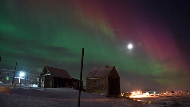Purple and green northern lights hang like curtains in the night sky over two wooden shacks in Nome.