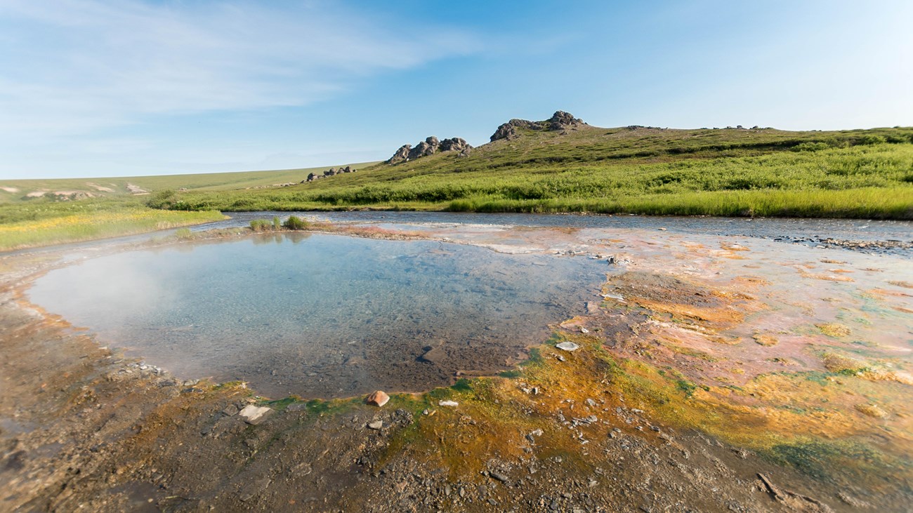 A shallow hot spring in a remote landscape is surrounded by rolling hills. 