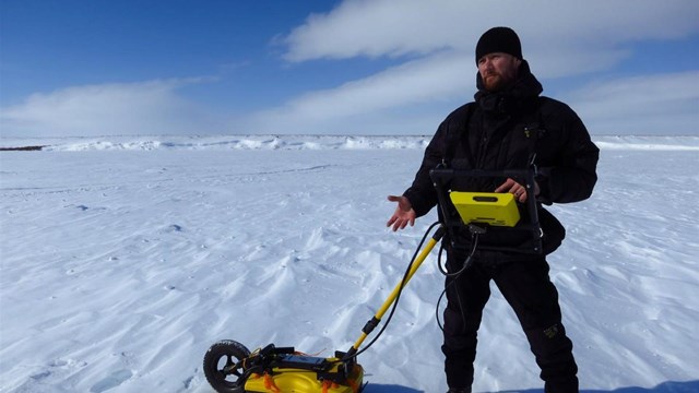 A scientist uses survey equipment while standing on the frozen tundra.