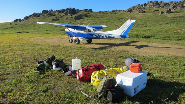 A small plane is parked at the end of a gravel air strip. A pile of gear is near the airstrip.
