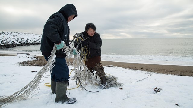 A man and woman work together to haul discarded fishing nets off a snowy beach.