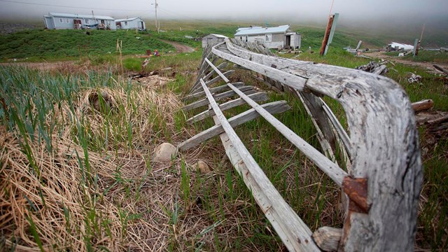 A frame of a traditional umiak with three houses in the background. 