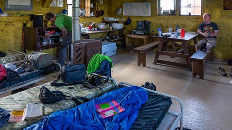 Two men in small room with yellow walls, several cots, and cooking paraphernalia.
